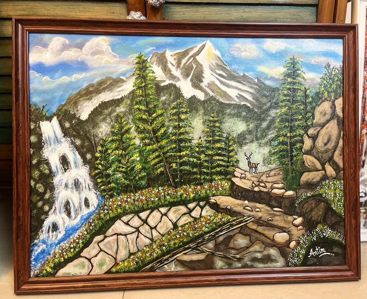 Snow-topped Himalayan mountain above a pine forest, waterfall, rocky stream, flowers, and a deer, in a brown frame.